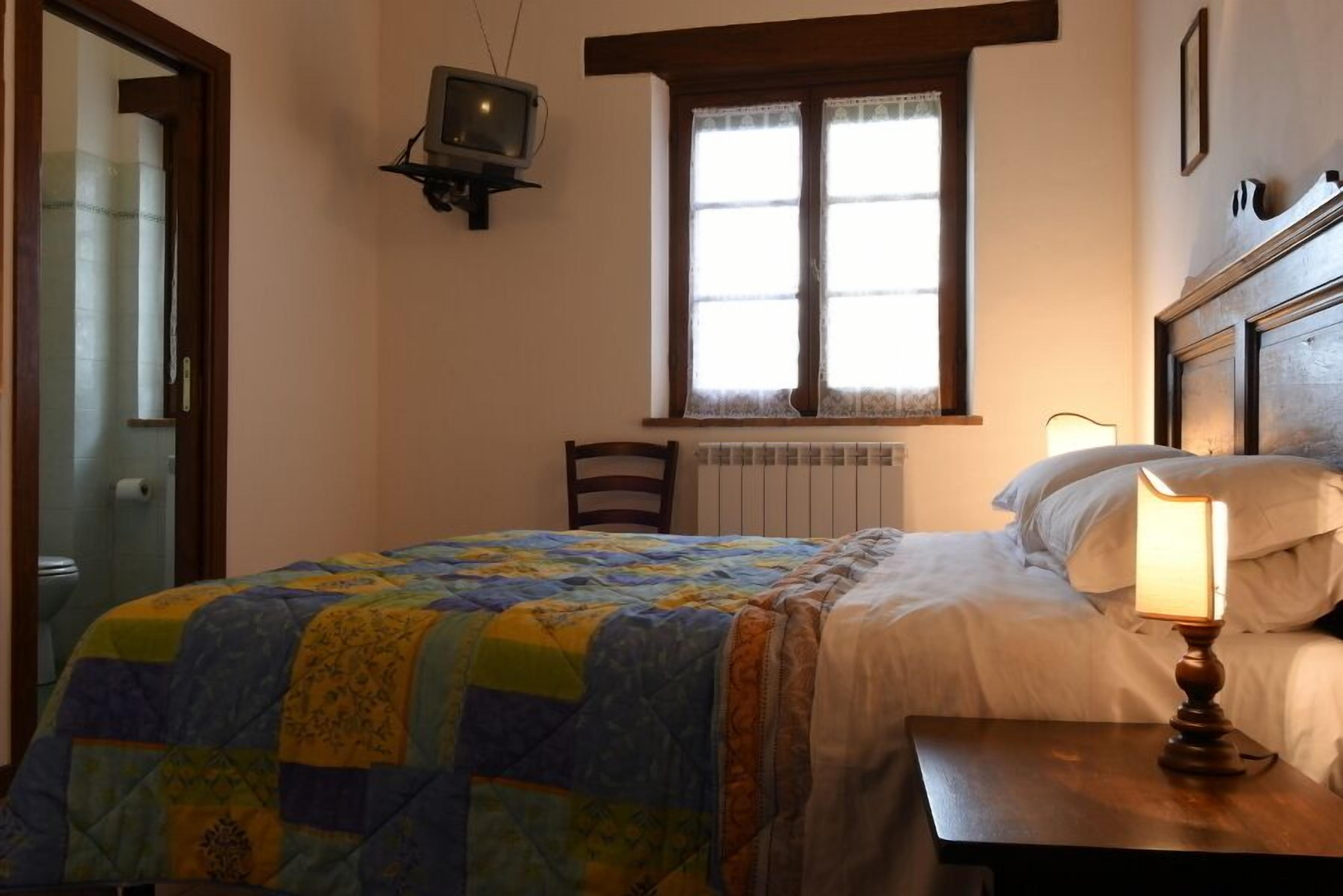 A bedroom with a bed, a nightstand with a lamp, a chair, a window with curtains, and a television mounted on the wall.