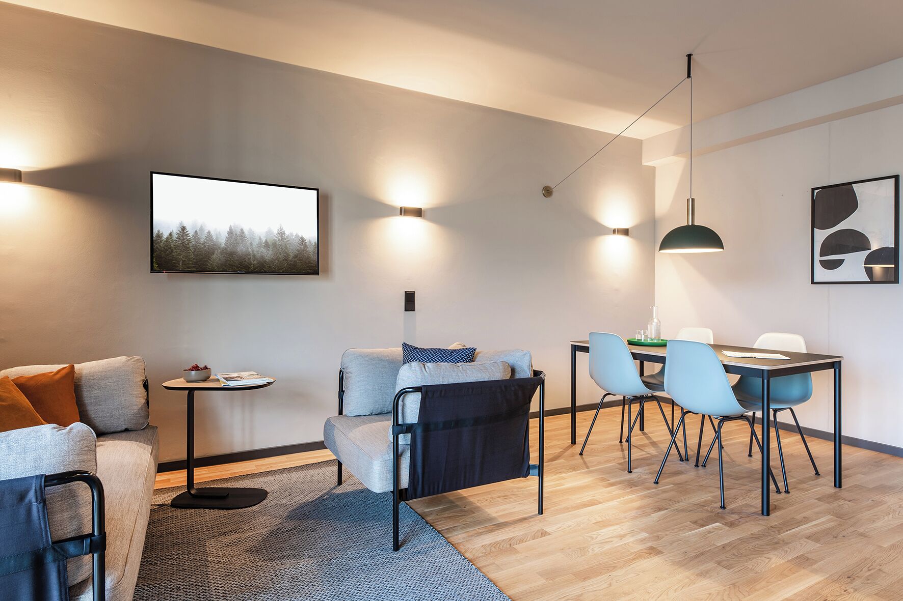 A modern living room with a sofa, a dining table with chairs, and a coffee table. There are wall-mounted lights and a painting on the wall.