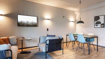 A modern living room with a sofa, a dining table with chairs, and a coffee table. There are wall-mounted lights and a painting on the wall.