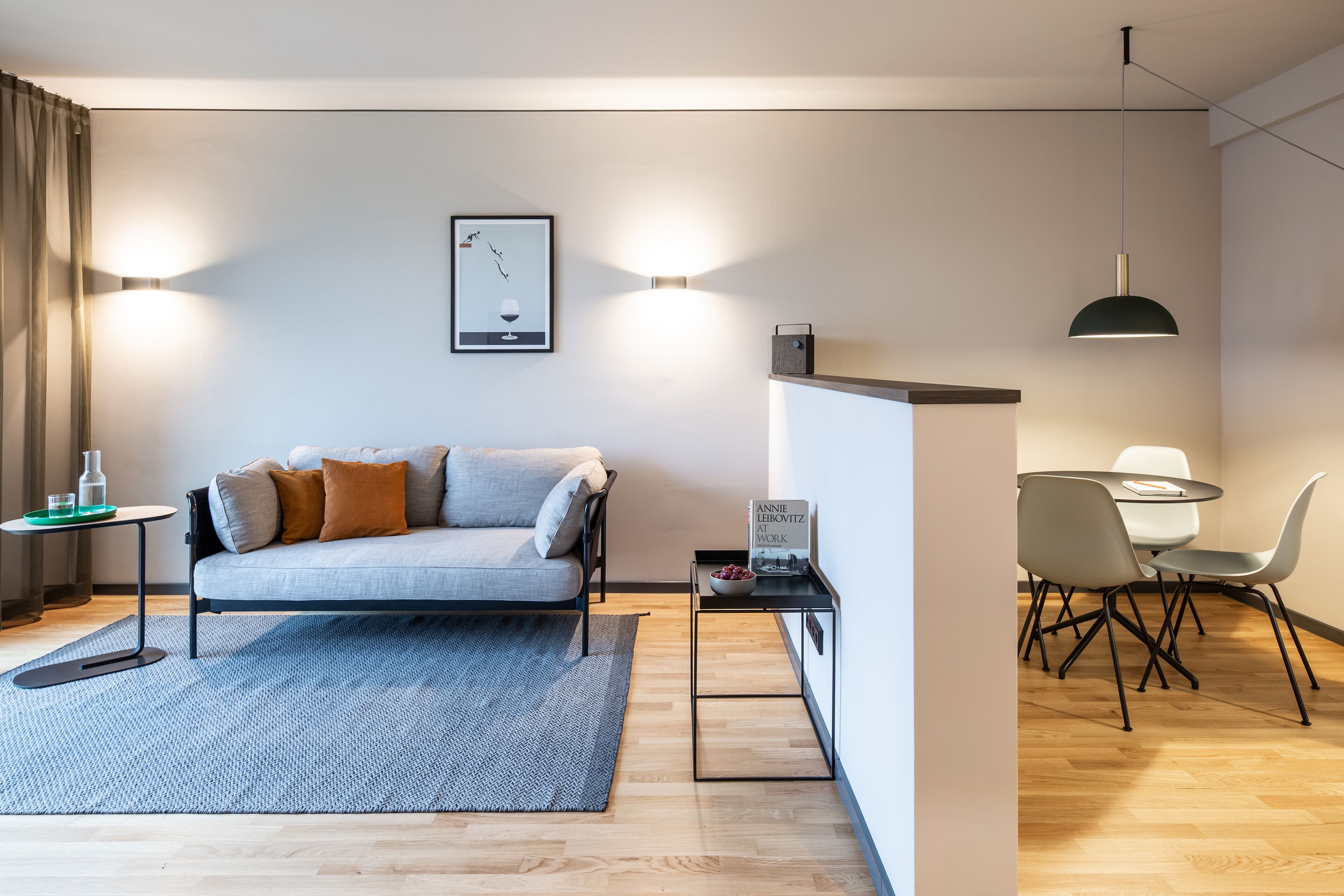 Een moderne woonkamer met een bank, een eethoek met stoelen, een klein tafeltje en een wandlamp.
