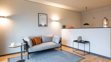 A modern living room with a grey sofa, a small round table with a cup and book, a grey rug, and a wall-mounted light fixture.