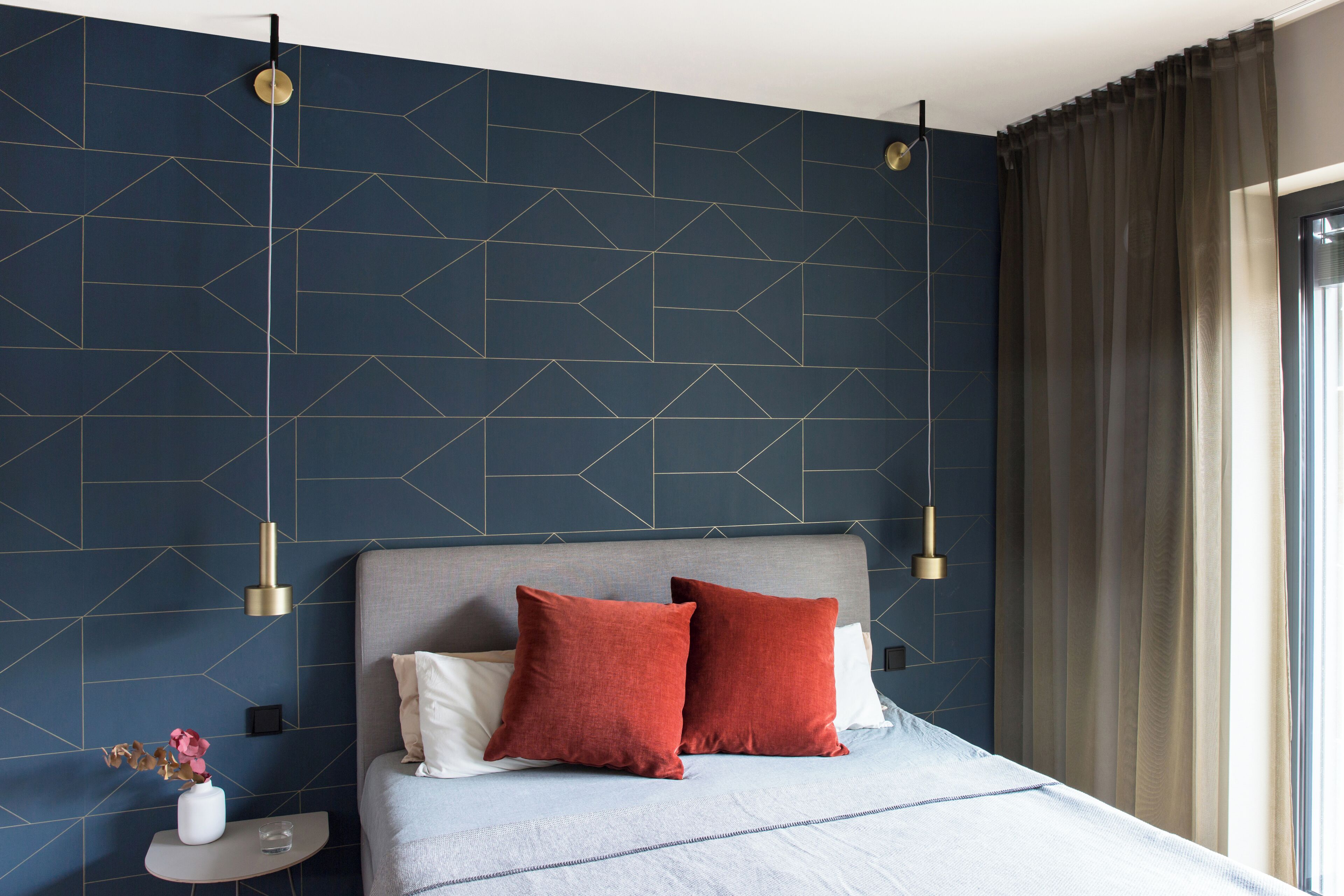 A modern bedroom with a geometric patterned wall, a grey upholstered headboard, and two red pillows.