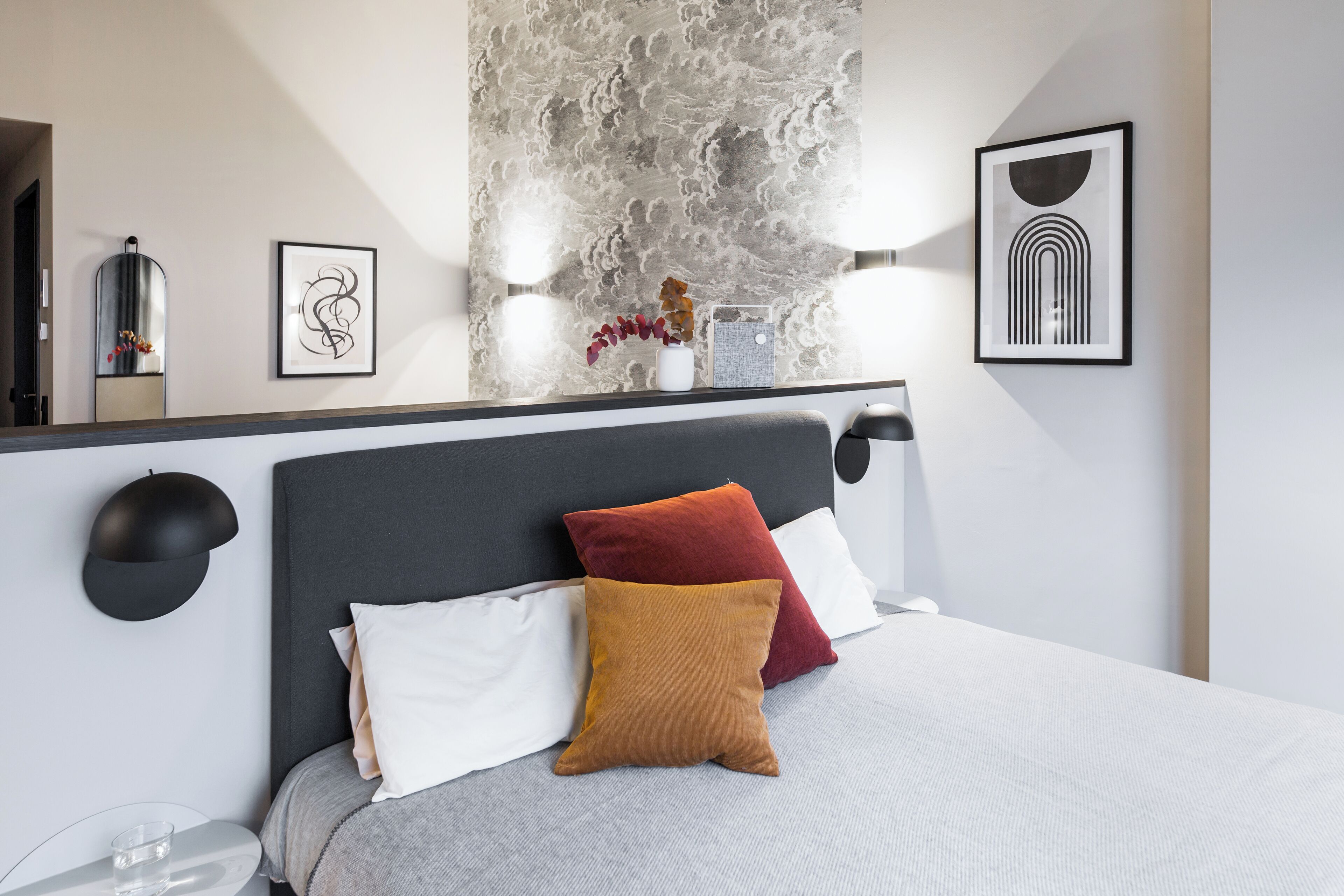 Un dormitorio moderno con cabecera gris, almohadas blancas y marrones, edredón gris y un espejo fijado a la pared.