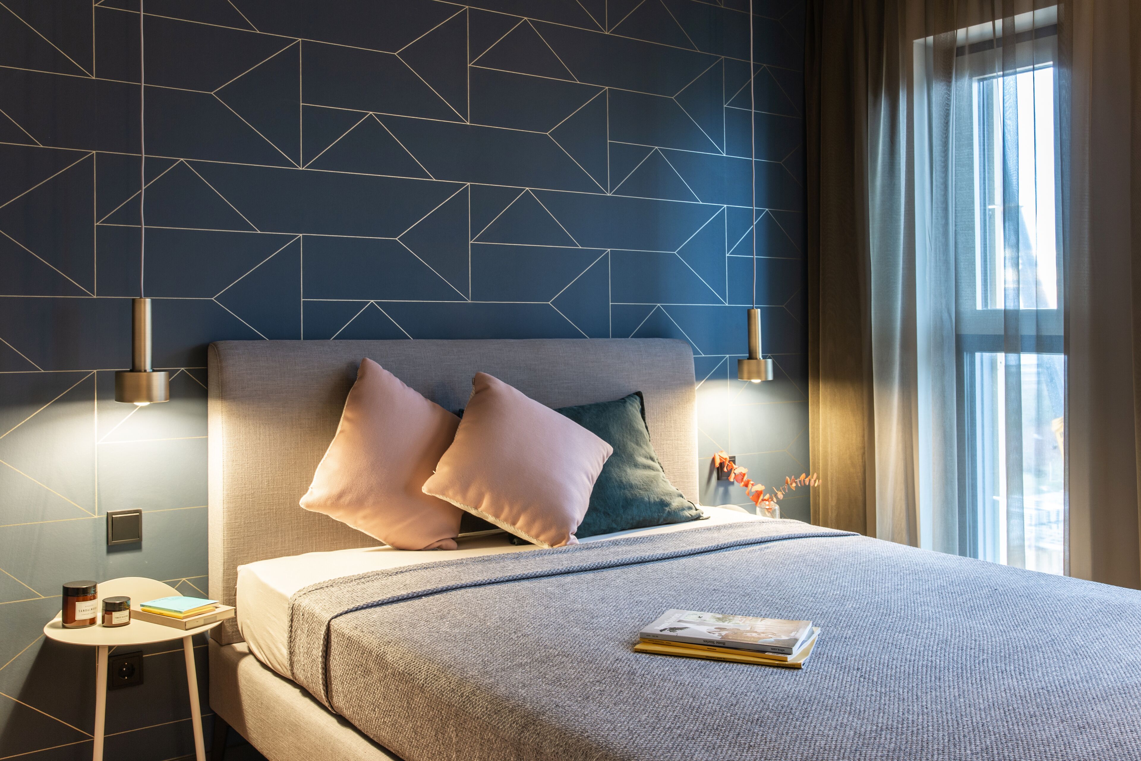 Une chambre d’hôtel moderne avec un mur orné d’un motif géométrique, un grand lit doté d’une tête de lit grise, deux lampes de chevet et une vue sur la ville par la fenêtre.