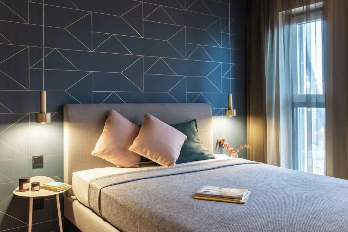 A modern hotel room with a geometric-patterned wall, a large bed with a grey headboard, two bedside lamps, and a view of the city through a window.