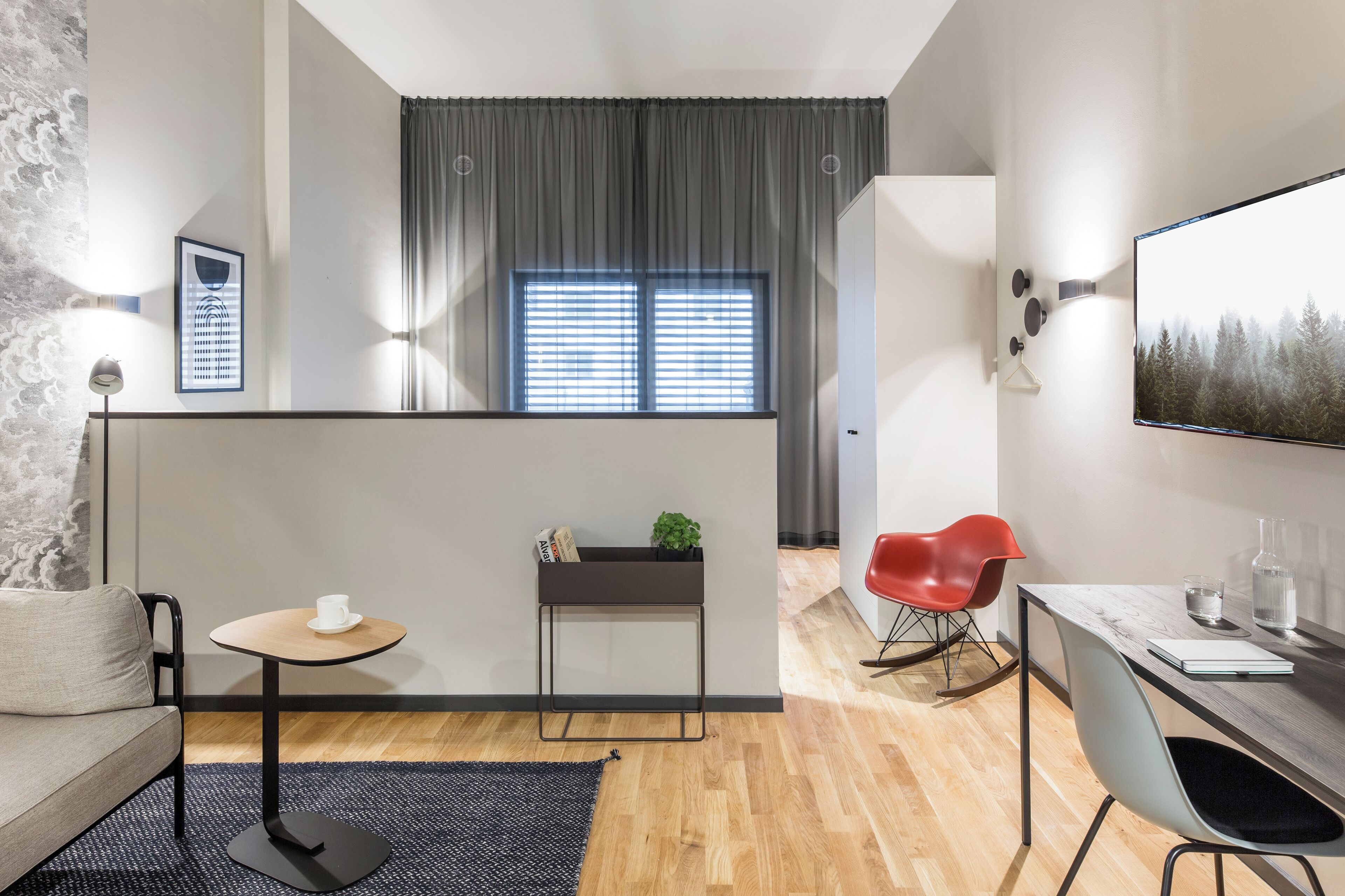 Een moderne woonkamer met een bank, een klein rond tafeltje, een rode stoel, een bureau met stoel en een groot raam met jaloezieën.