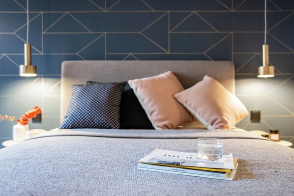 Una cama bien hecha con un cabecero gris, dos almohadas, y una pila de libros junto con un vaso de agua sobre la cama.