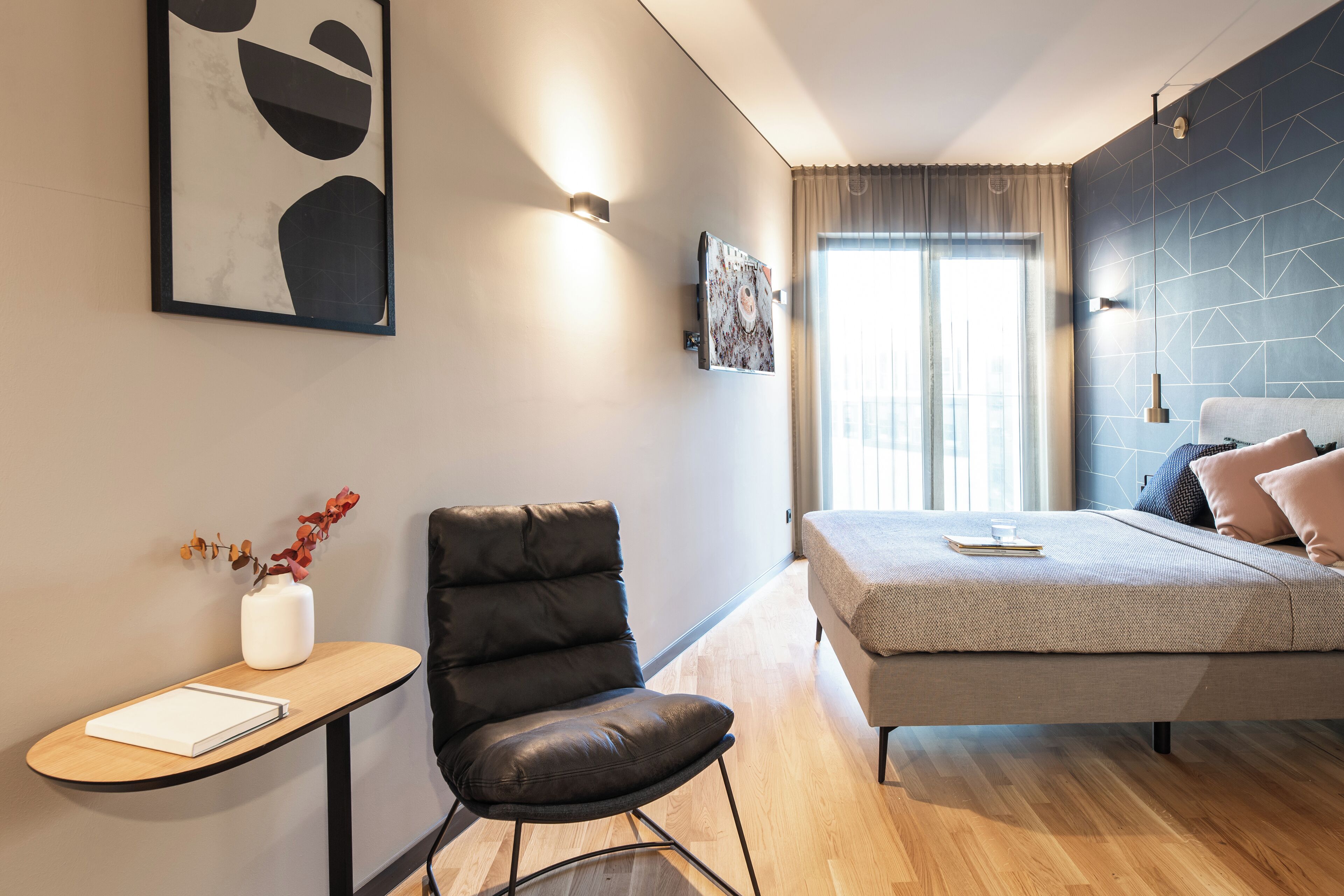 Un dormitorio moderno con cama, una silla, una mesita y un jarrón con flores.