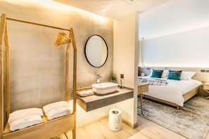 A modern hotel room with a wooden bath chair, a large mirror, a white sink, a bedside table, a bed with a grey headboard, and a patterned rug. - Suzhou BuerJu (Suzhou)