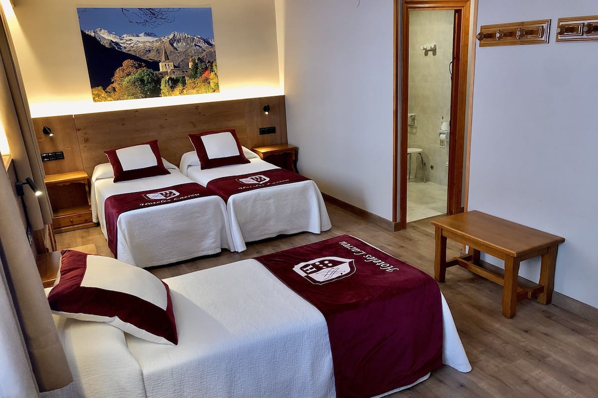 Ein Hotelzimmer mit zwei Betten, einem Holzkopfende, einem Bergbild, einer Holzbank und einer Badezimmertür.