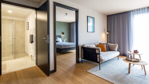 A modern hotel room with a glass shower, a bed with a grey headboard, a sofa with cushions, a small round table with a bottle and a bowl, and a large window with curtains. - ipartment Boeblingen @HOME by Best Western (Boeblingen)