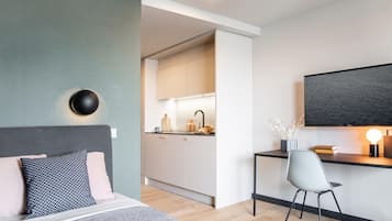 A modern hotel room with a bed, a desk with a chair, a TV, and a kitchenette.