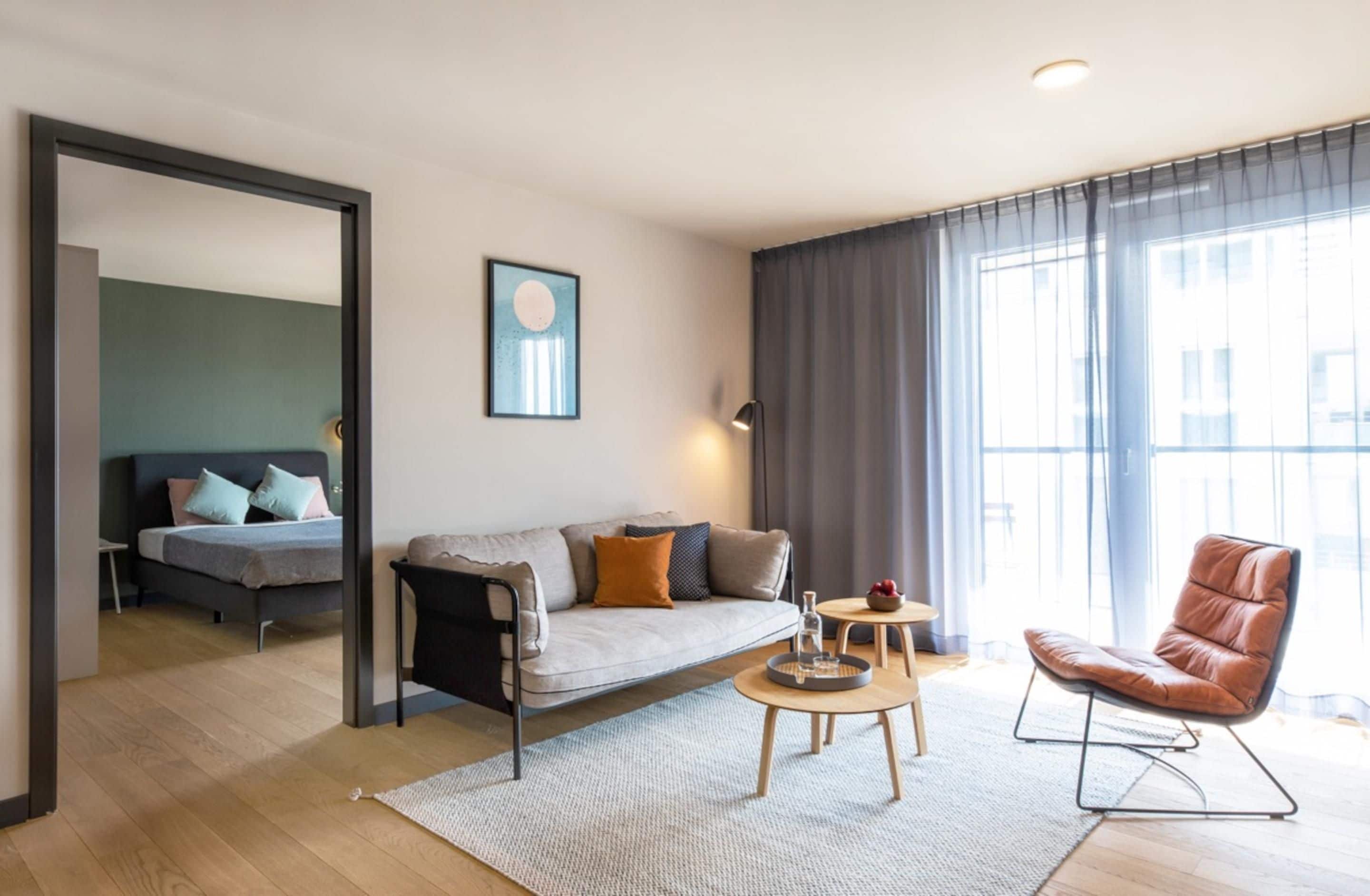 Una sala de estar moderna con sofá, mesa de centro y sillón, una cama al fondo, un ventanal con cortinas y un cuadro en la pared.