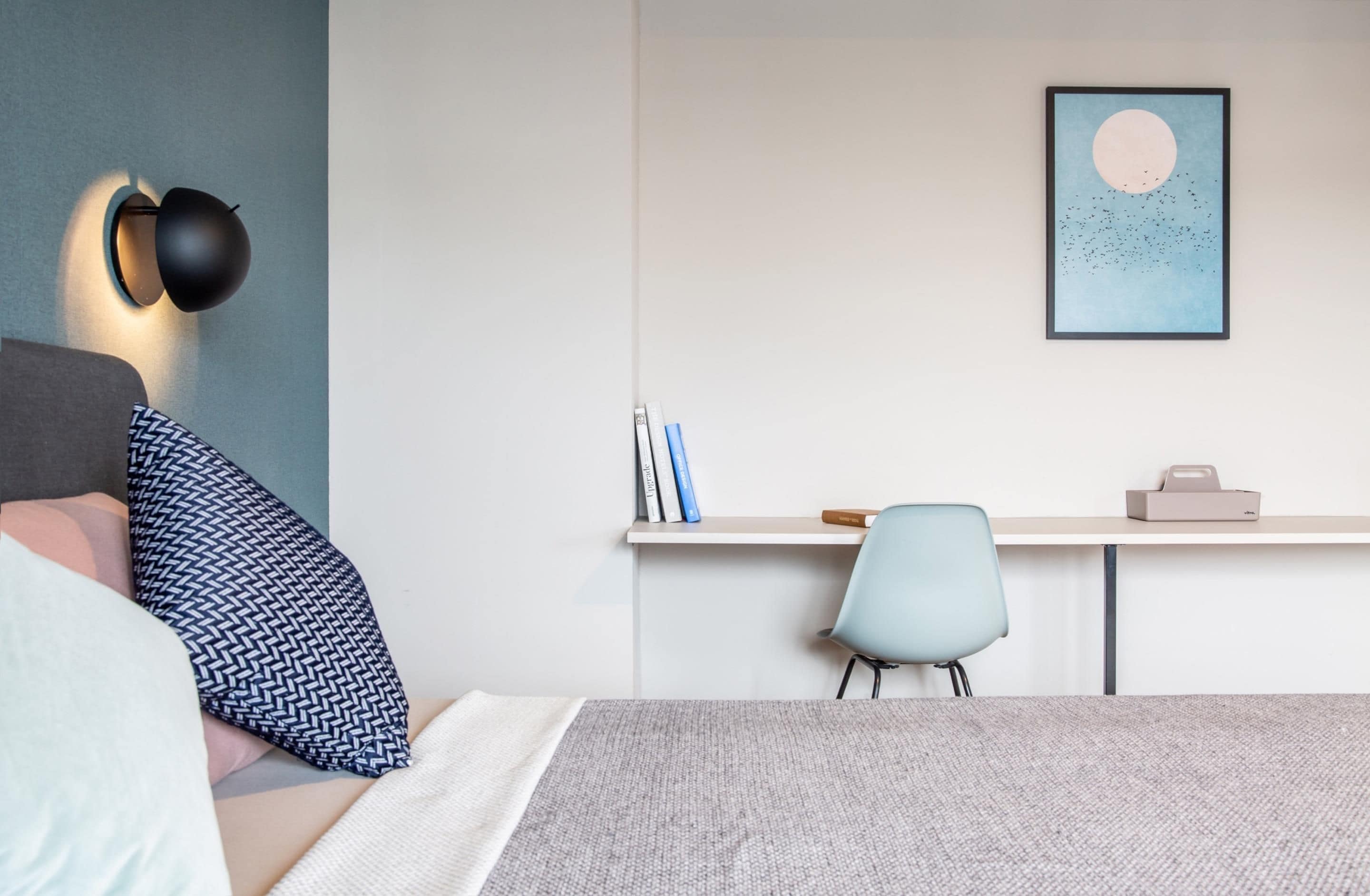 A bedroom with a bed, a desk, a chair, a wall-mounted lamp, and a framed picture.