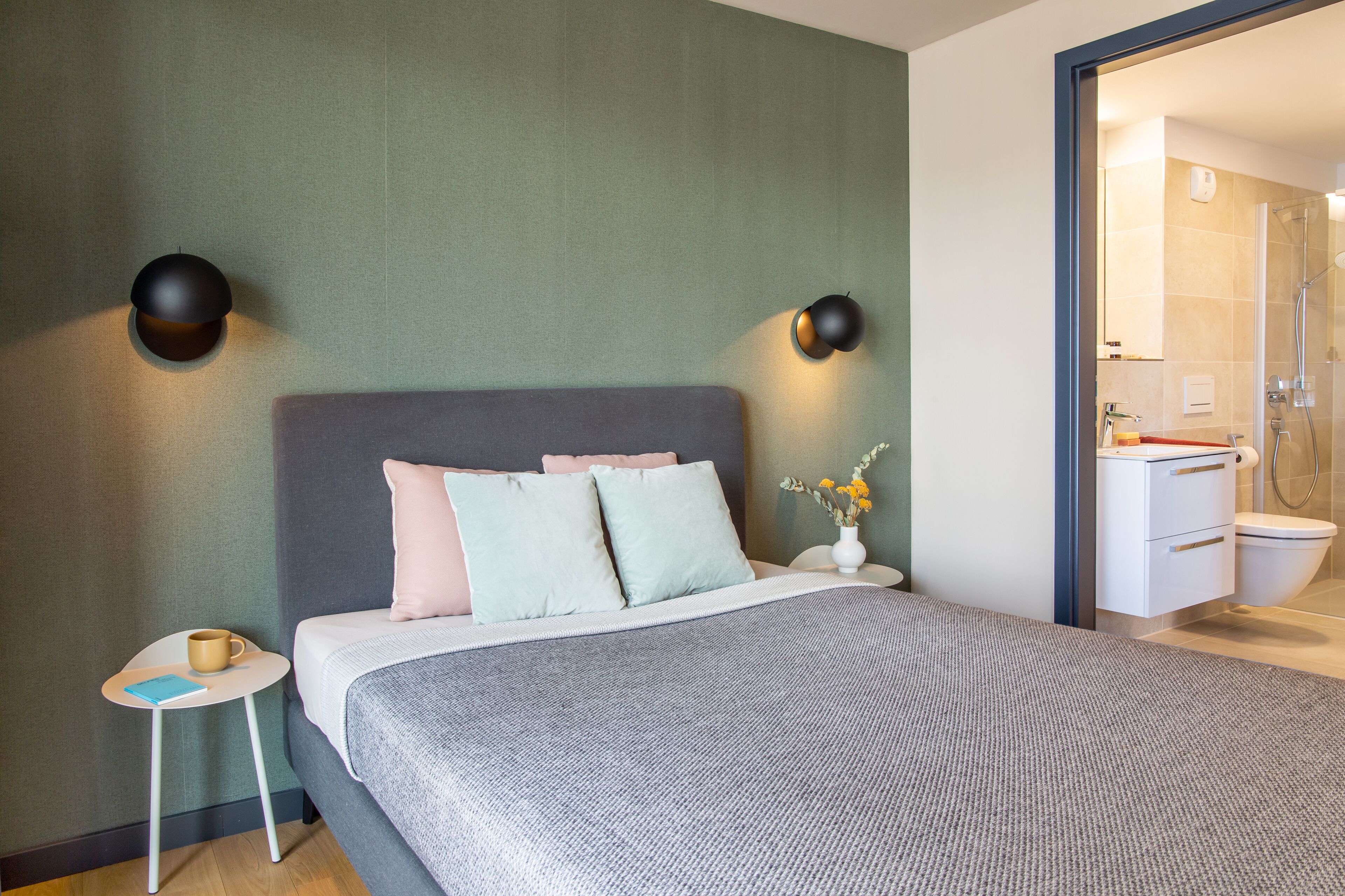 A modern hotel room with a grey bed, a small bedside table with a cup and a book, and a vase with flowers on the nightstand.