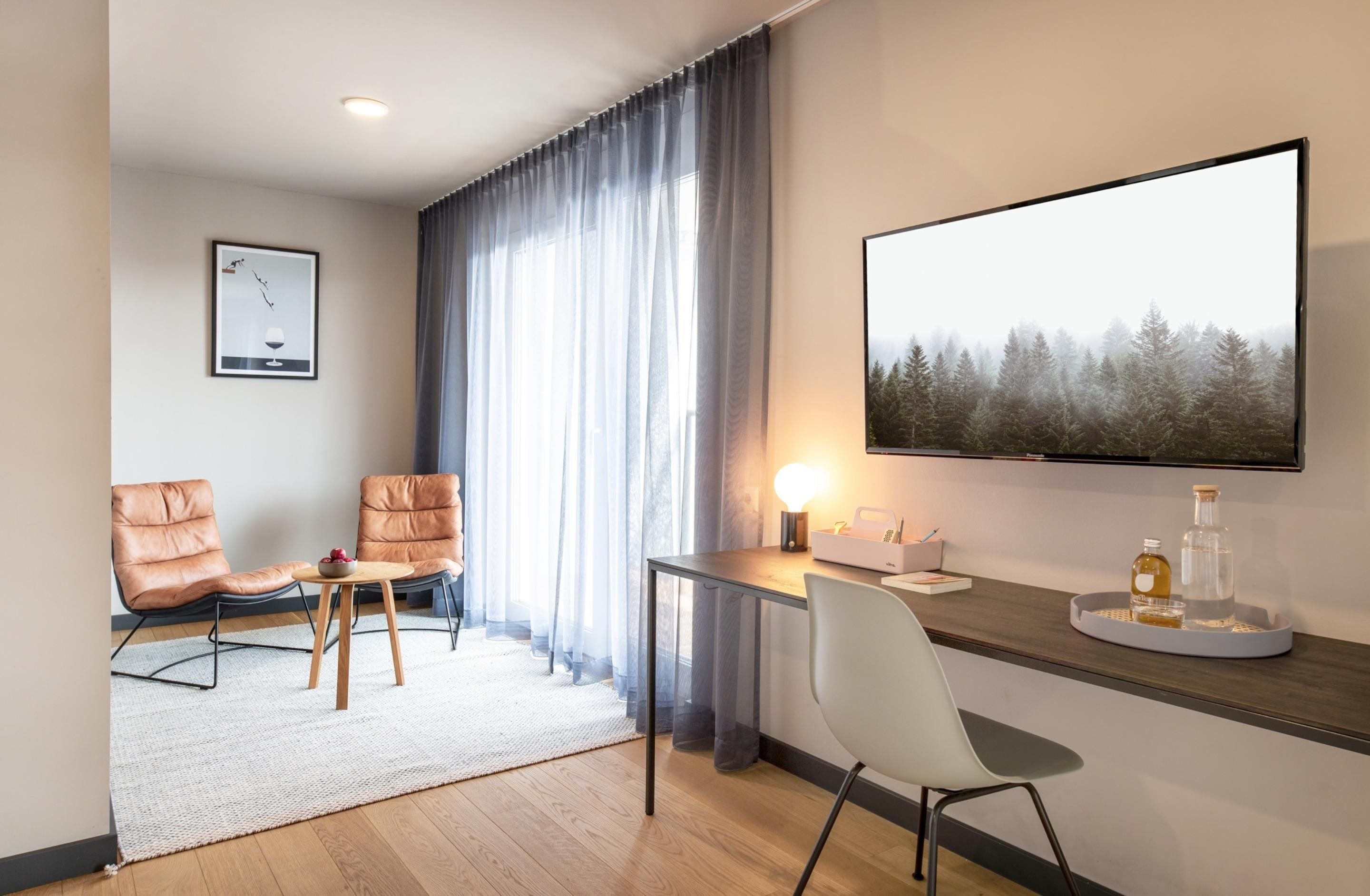 Una habitación de hotel moderna con un televisor de pantalla plana grande, un escritorio con lámpara, dos sillones de cuero, una mesita y un cuadro enmarcado en la pared.