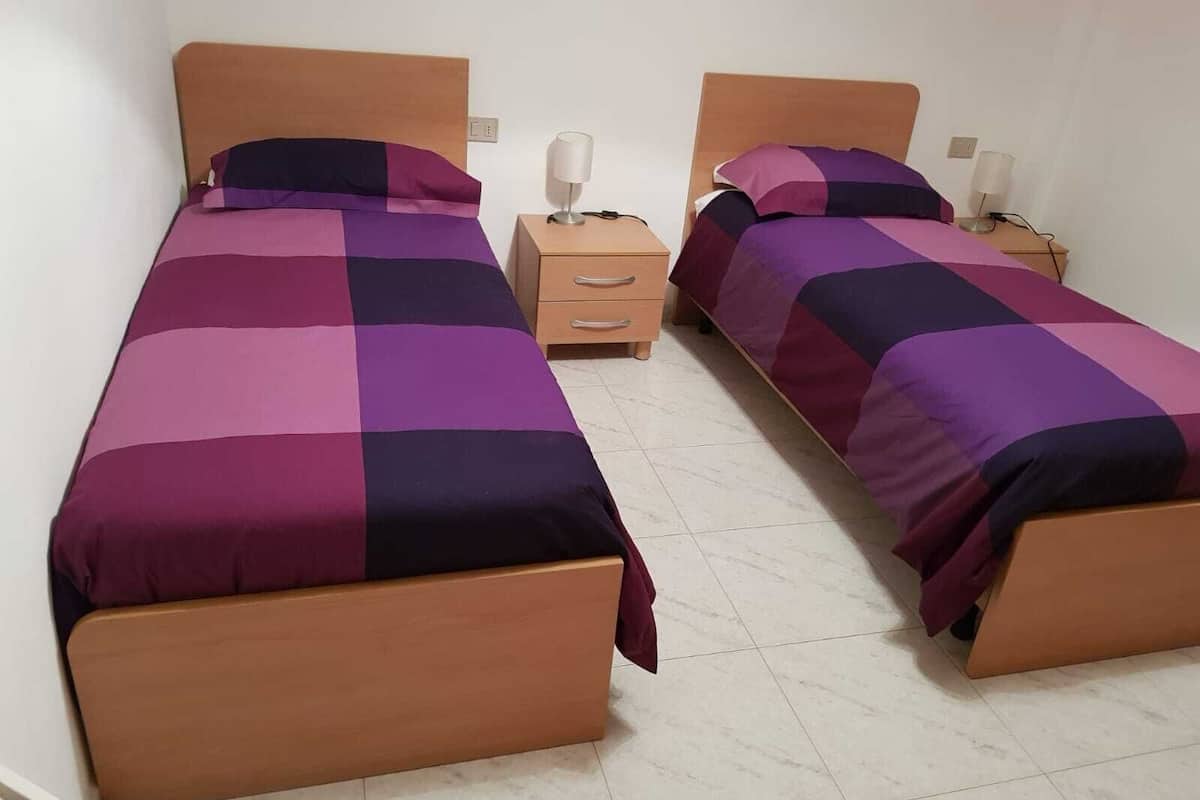 A room with two beds, each with a wooden headboard and a purple and black checkered bedspread. There are bedside tables with lamps and a small wooden drawer unit between the beds.