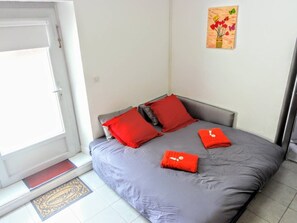 A bedroom with a grey bed, red pillows, a small red towel, and a painting of flowers on the wall.