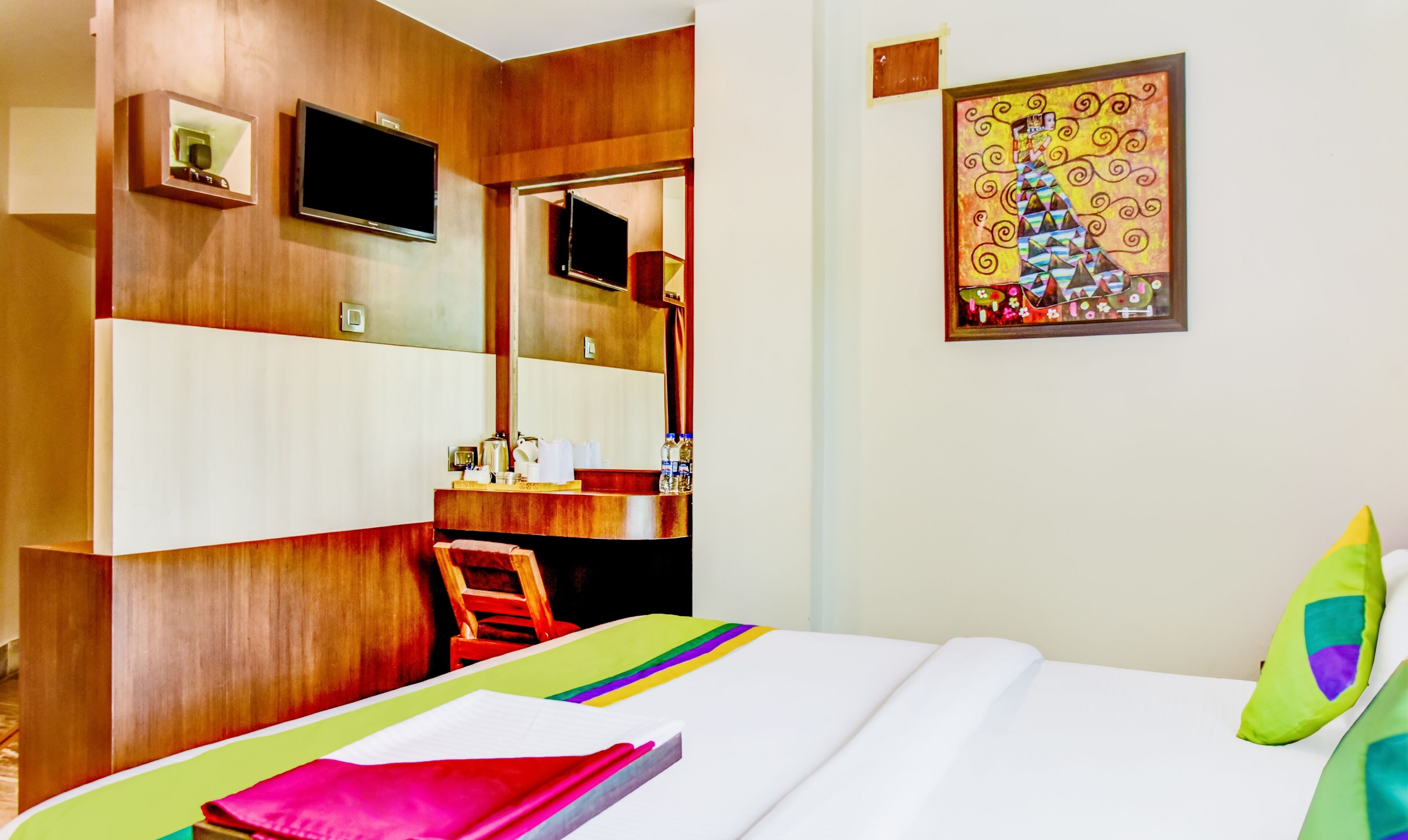 A hotel room with a bed, a desk, a chair, a TV, and a painting on the wall.