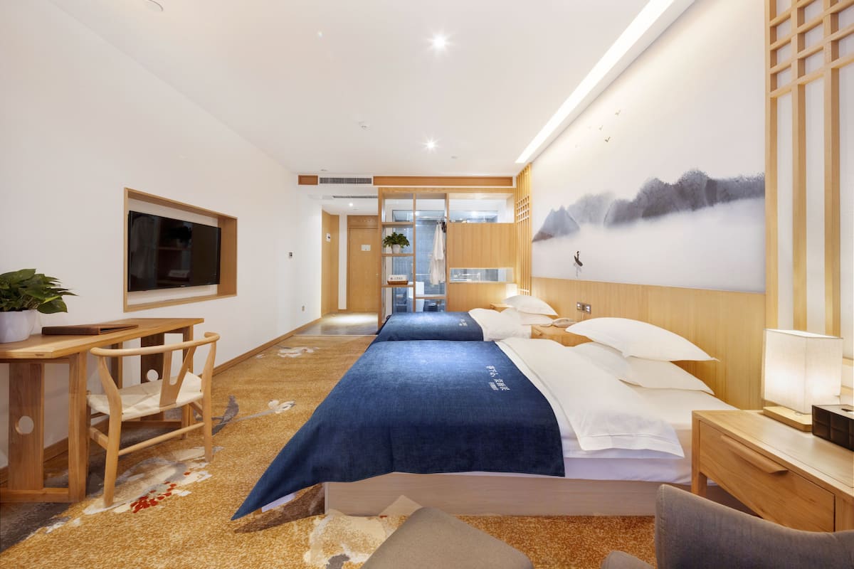 A hotel room with a bed, bedside table, lamp, TV, and a view of a mountain landscape.