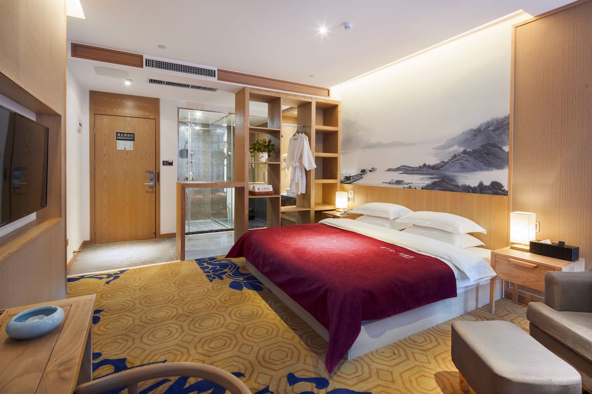 A hotel room with a large bed, a wooden wardrobe, a desk with a chair, a small table with a bowl, and a painting on the wall.