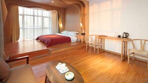 A modern hotel room with a wooden floor, a bed with red bedding, a wooden desk with a kettle, and a chair.