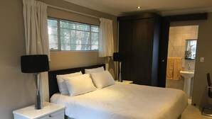 A neatly made bed with white linens, two bedside tables with lamps, a window with blinds, and a bathroom visible in the background. - Northwold Comfort Living (Randburg)