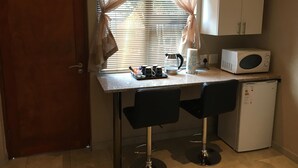 A compact kitchenette with a microwave, coffee maker, and a small refrigerator. There are two black bar stools and a window with sheer curtains. - Northwold Comfort Living (Randburg)