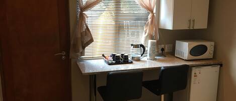 A compact kitchenette with a microwave, coffee maker, and a small refrigerator. There are two black bar stools and a window with sheer curtains.