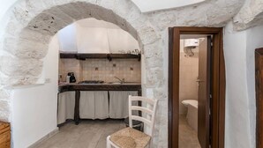 A rustic kitchen with a stone archway, a wooden chair, and a view into the adjacent bathroom. - Trullieu Guesthouse (Alberobello)