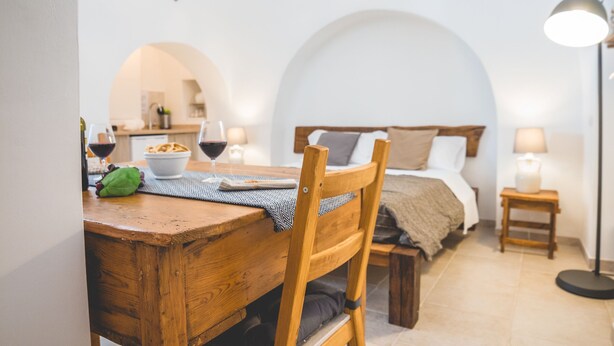 A cozy room with a wooden dining table, a bed with a wooden headboard, and a small bedside table with a lamp. - Trullieu Guesthouse (Alberobello)