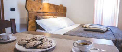 A neatly arranged breakfast table with a plate of cookies and a cup of coffee, a wooden headboard, and a bed with a blue blanket and pillows in the background.