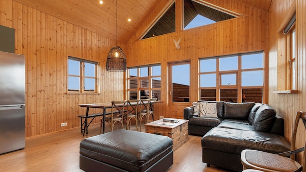 A spacious living area with a wooden interior, a leather sofa, a dining table, and a large window providing natural light. - Ocean Break Cabins (Suðurnesjabær)