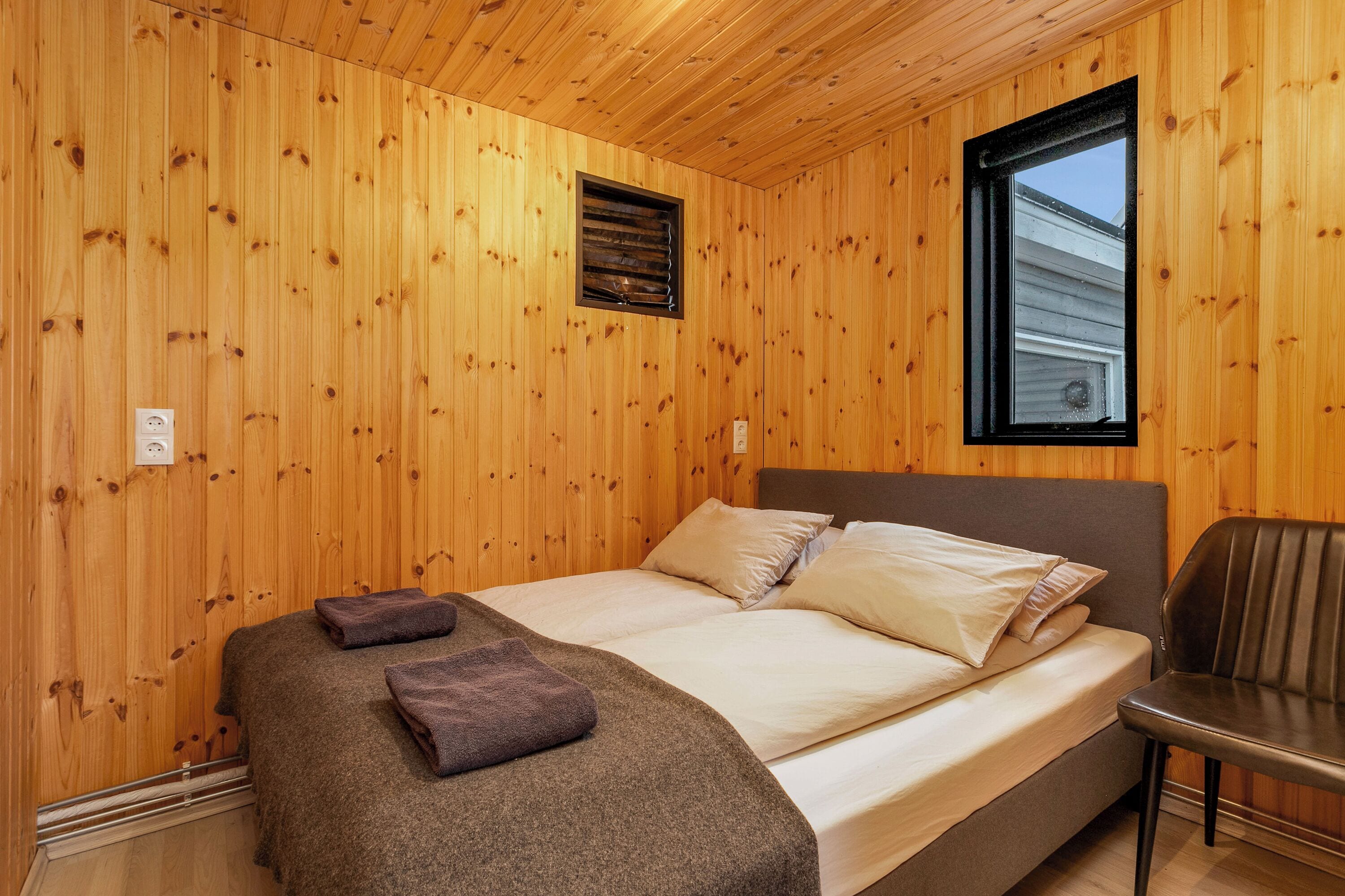 Een kamer met houten lambrisering, een bed, kussens, een stoel en een raam.