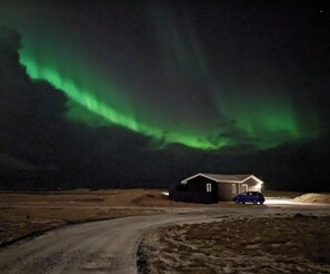 A house with a dirt road leading up to it, under a night sky with green aurora borealis. - Ocean Break Cabins (Suðurnesjabær)