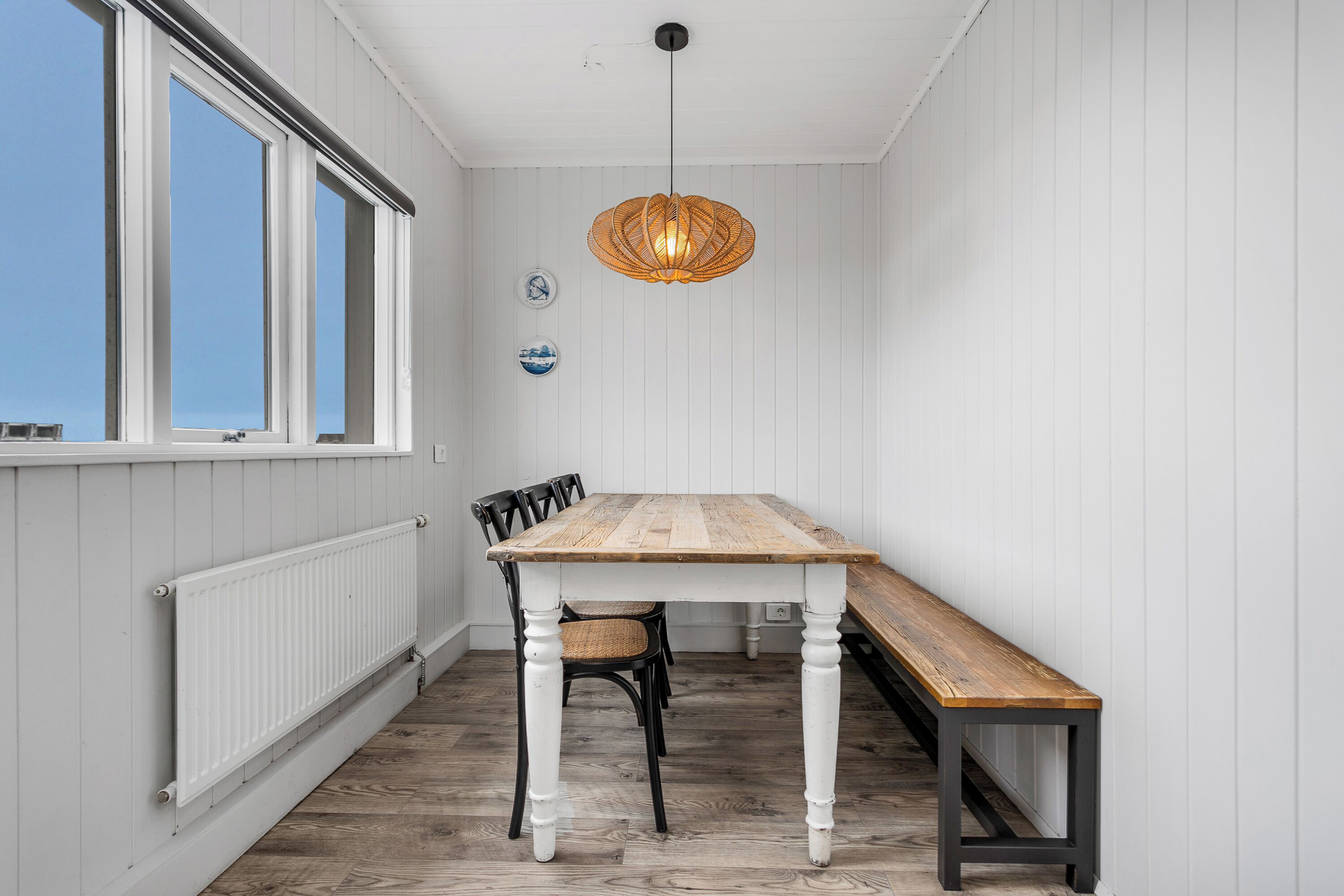 Un coin repas avec une table et des bancs en bois, des murs recouverts de panneaux blancs, une fenêtre et un luminaire suspendu.