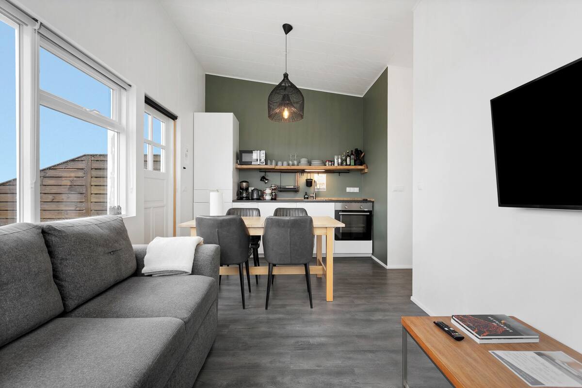 Una sala de estar moderna con un sofá gris, una mesa de comedor con sillas, una zona de cocina con microondas y un televisor montado en la pared.