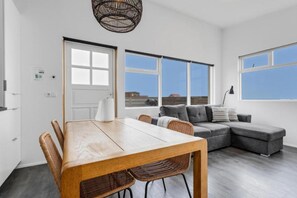 A modern living room with a wooden dining table, wicker chairs, a grey sectional sofa, and large windows offering a view of the city. - Ocean Break Cabins (Suðurnesjabær)