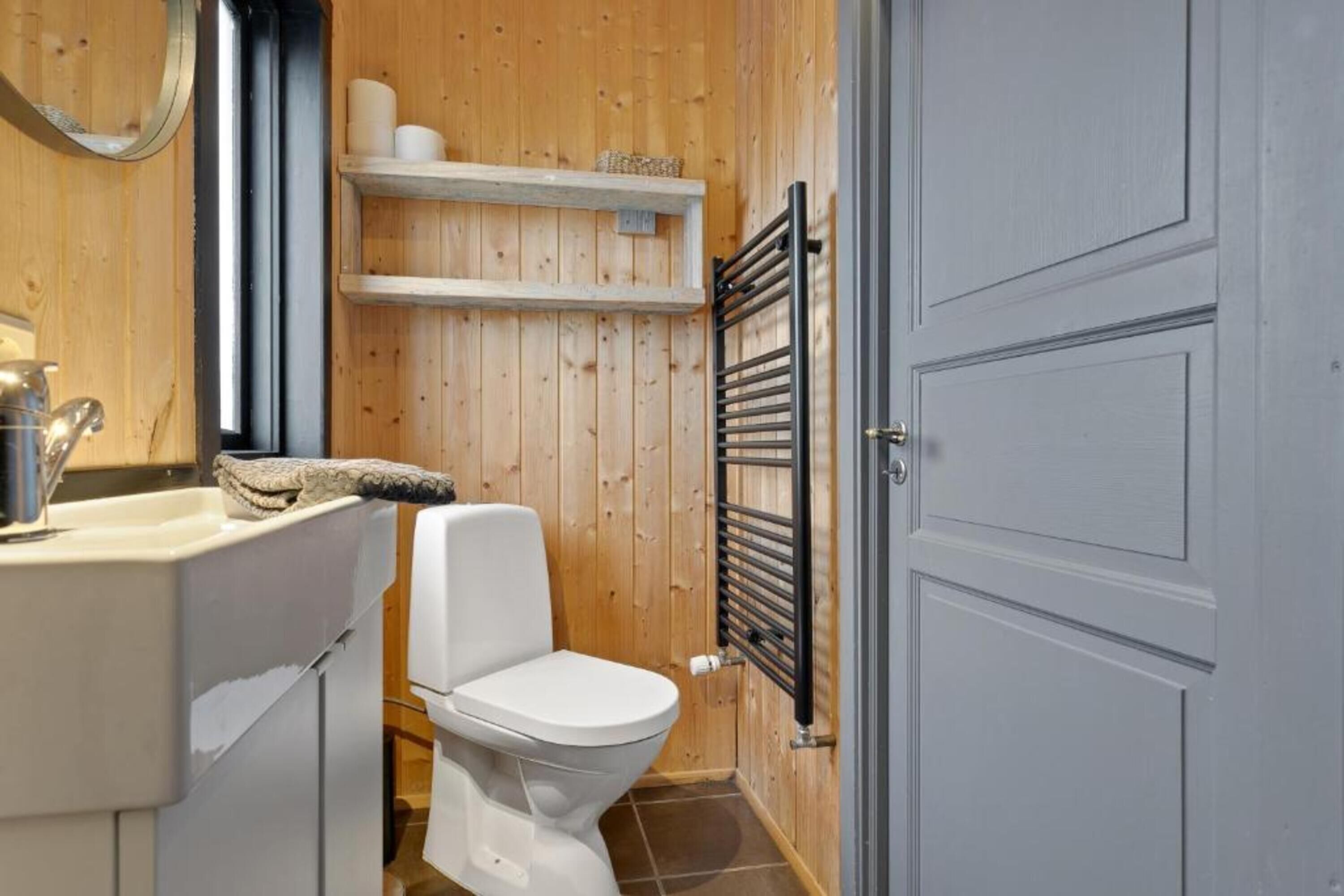 A compact bathroom with a white toilet, wooden walls, a small shelf with toiletries, and a towel rack.