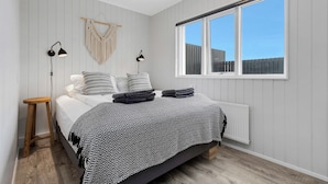 A bedroom with a bed, bedside lamps, a wooden stool, a macrame wall hanging, and a window with a view of a fence. - Ocean Break Cabins (Suðurnesjabær)
