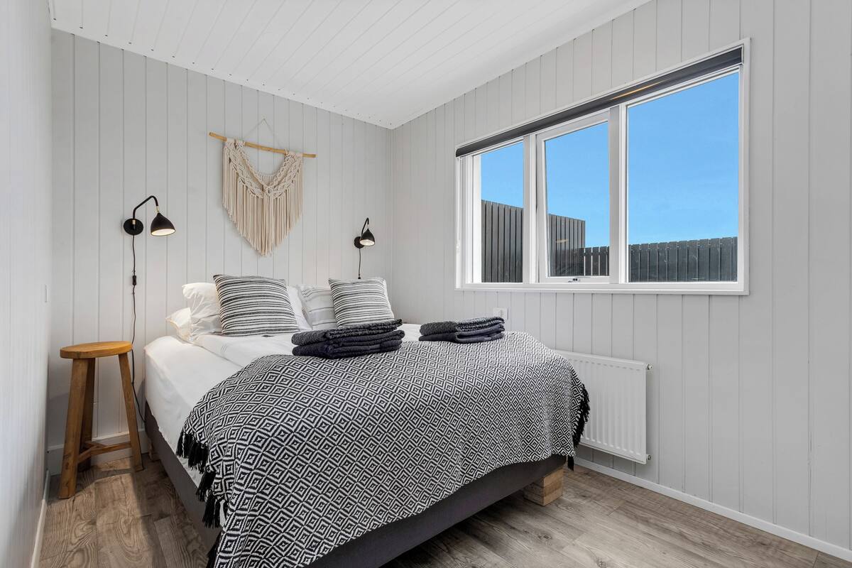 Un dormitorio con cama, lámparas de noche, un taburete de madera, un colgador de macramé y una ventana con vista a una cerca.
