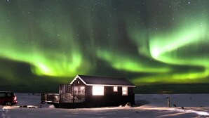 A cabin with a snow-covered ground and a starry night sky with aurora borealis. - Ocean Break Cabins (Suðurnesjabær)