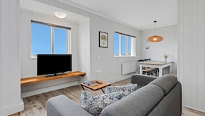 A modern living room with a grey sofa, a wooden coffee table, and a flat-screen TV mounted on a white wall. There is a dining area with a table and chairs, and a hanging pendant light. - Ocean Break Cabins (Suðurnesjabær)