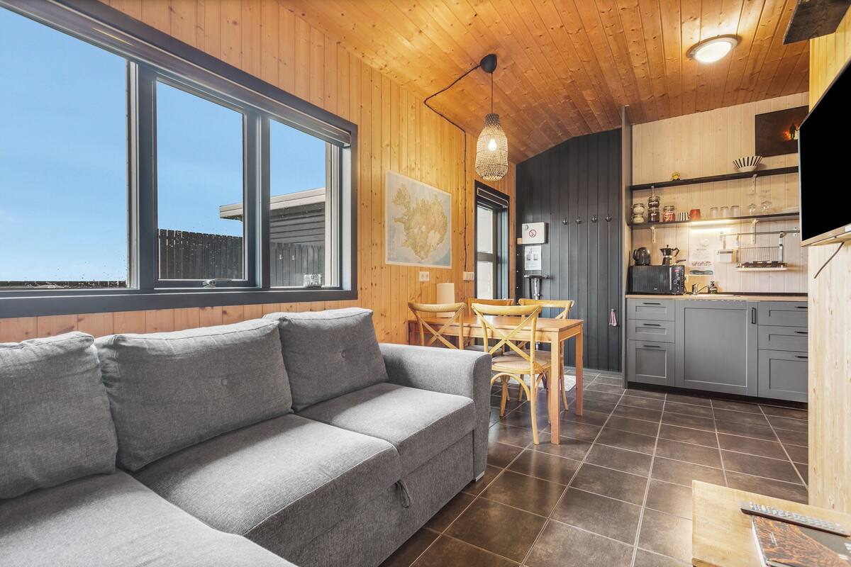 Un interior moderno con un sofá gris, techo de madera y una zona de cocina con armarios grises y una mesa de comedor de madera.