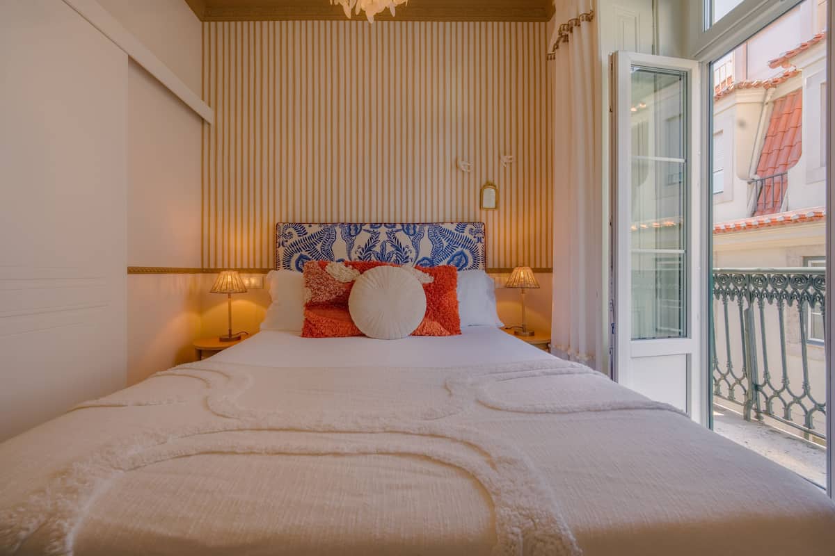 Une chambre d’hôtel avec un grand lit, un papier peint à rayures, une tête de lit décorative et un balcon doté d’une balustrade en fer forgé.