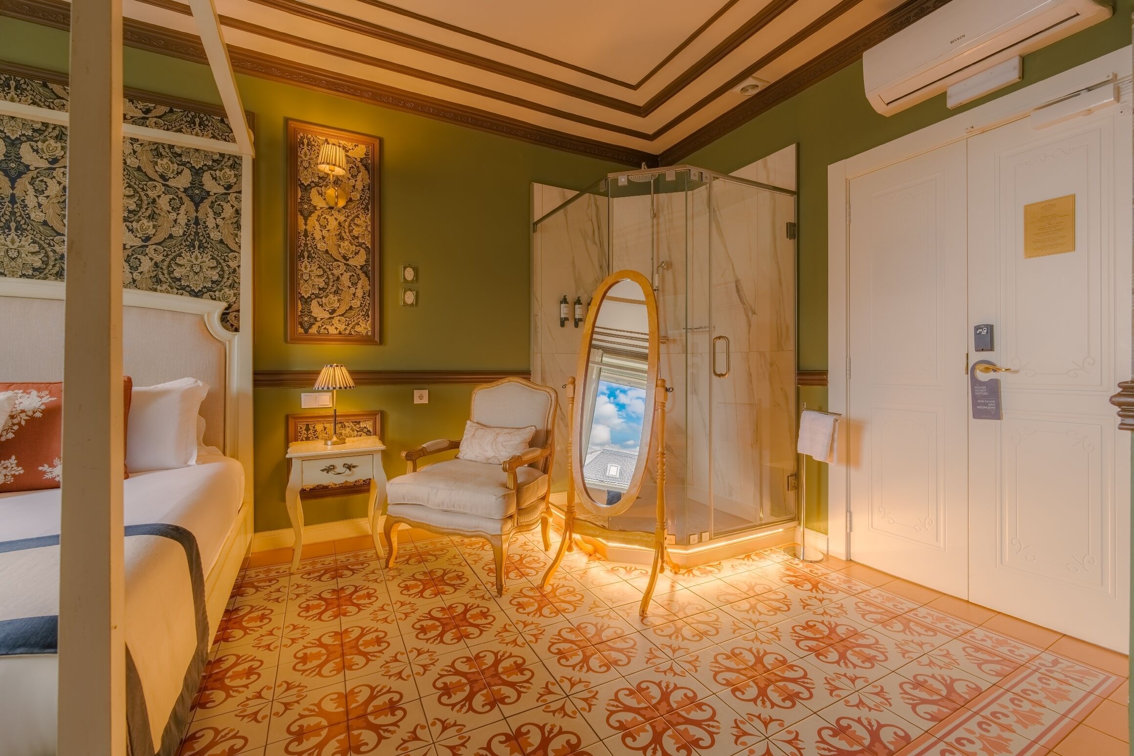 A room with a bed, a chair, a vanity with a mirror, a shower area, and a white door.