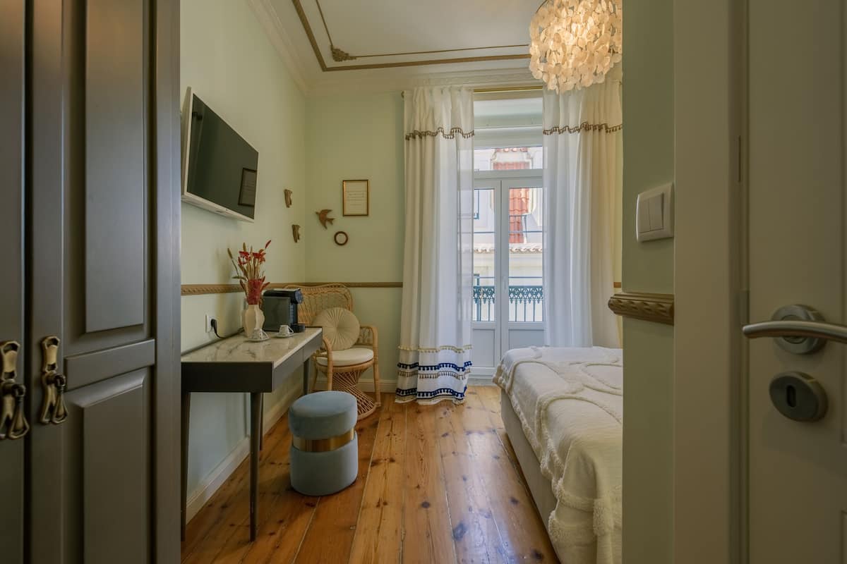 Une chambre avec un plancher en bois, un lit, un bureau avec une chaise, un petit tabouret, un téléviseur fixé au mur et une fenêtre avec des rideaux.
