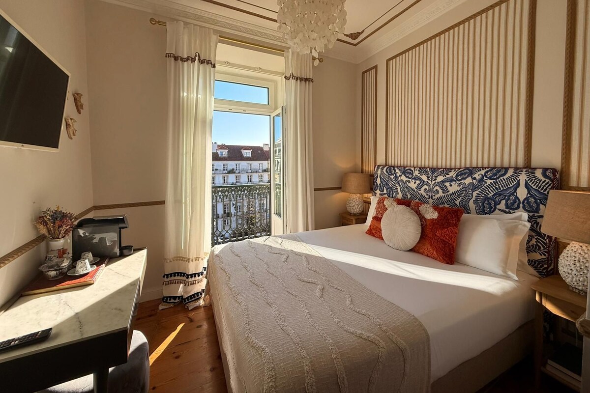 A hotel room with a large bed, a desk with a chair, a television, and a balcony with curtains.