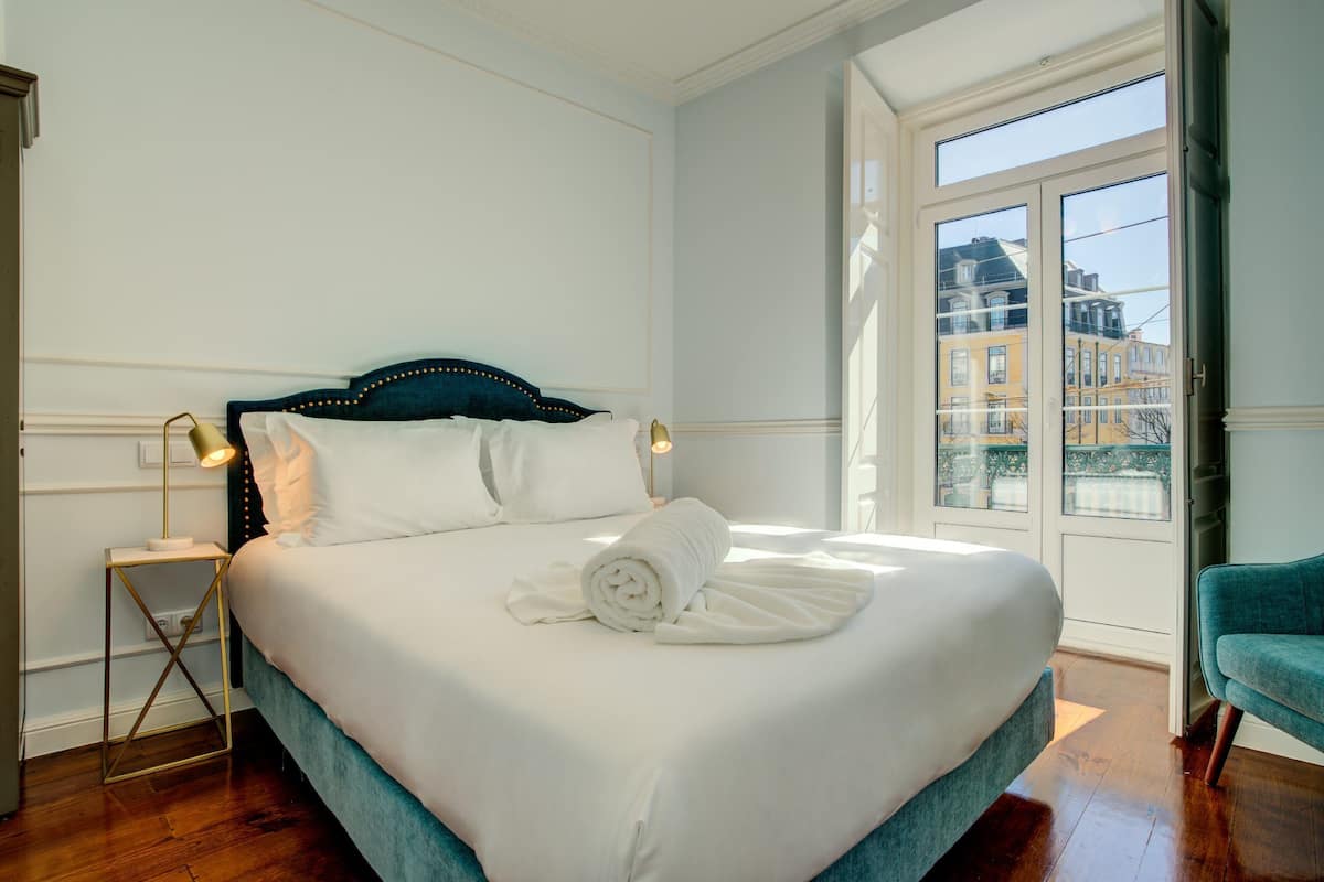 Un lit bien fait, avec du linge de lit blanc, une tête de lit sombre et une serviette enroulée posée sur le lit. Une petite table de chevet avec une lampe et une chaise se trouvent à droite.