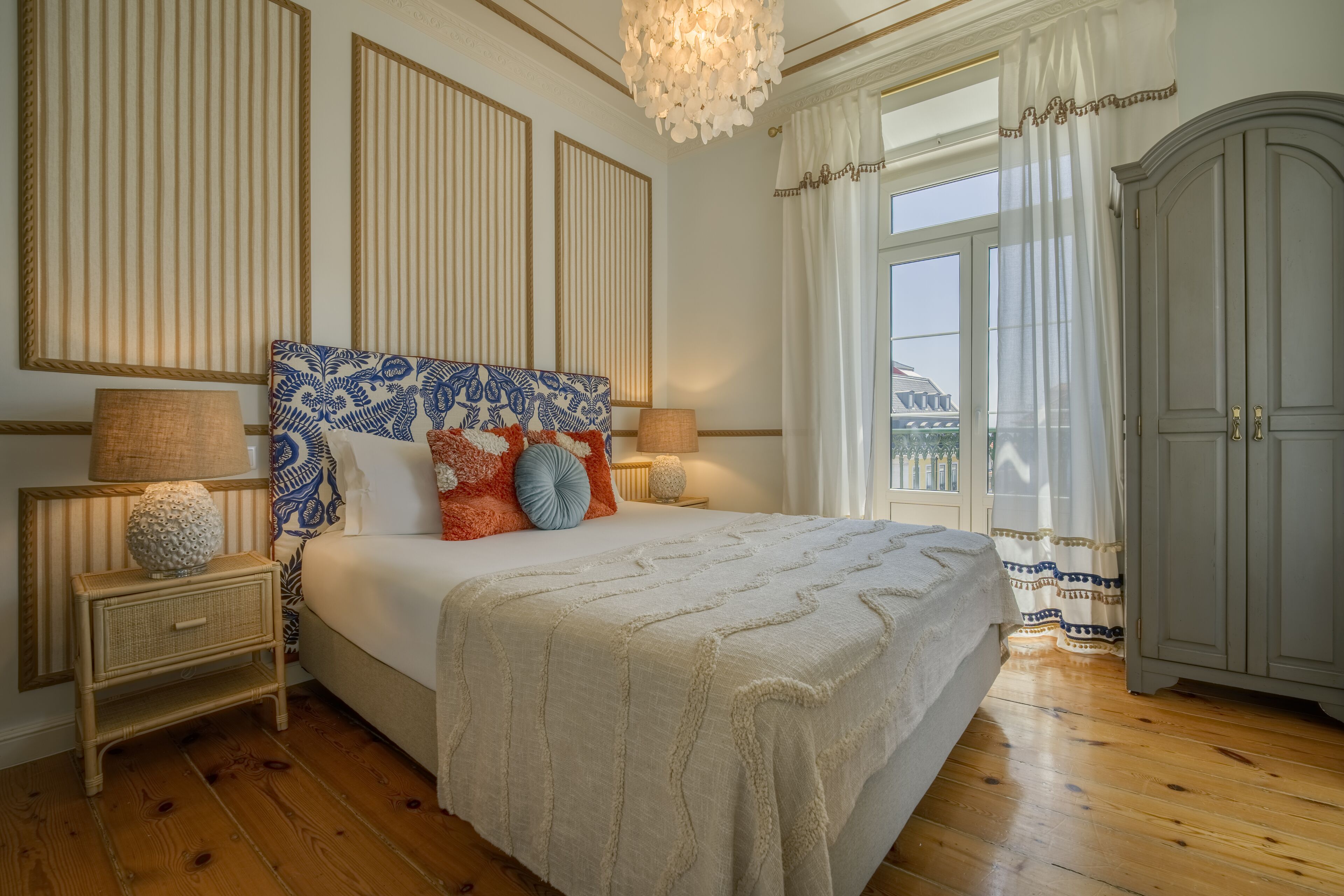 A bedroom with a large bed, two bedside tables, a nightstand, a chandelier, a wardrobe, and a window with curtains.